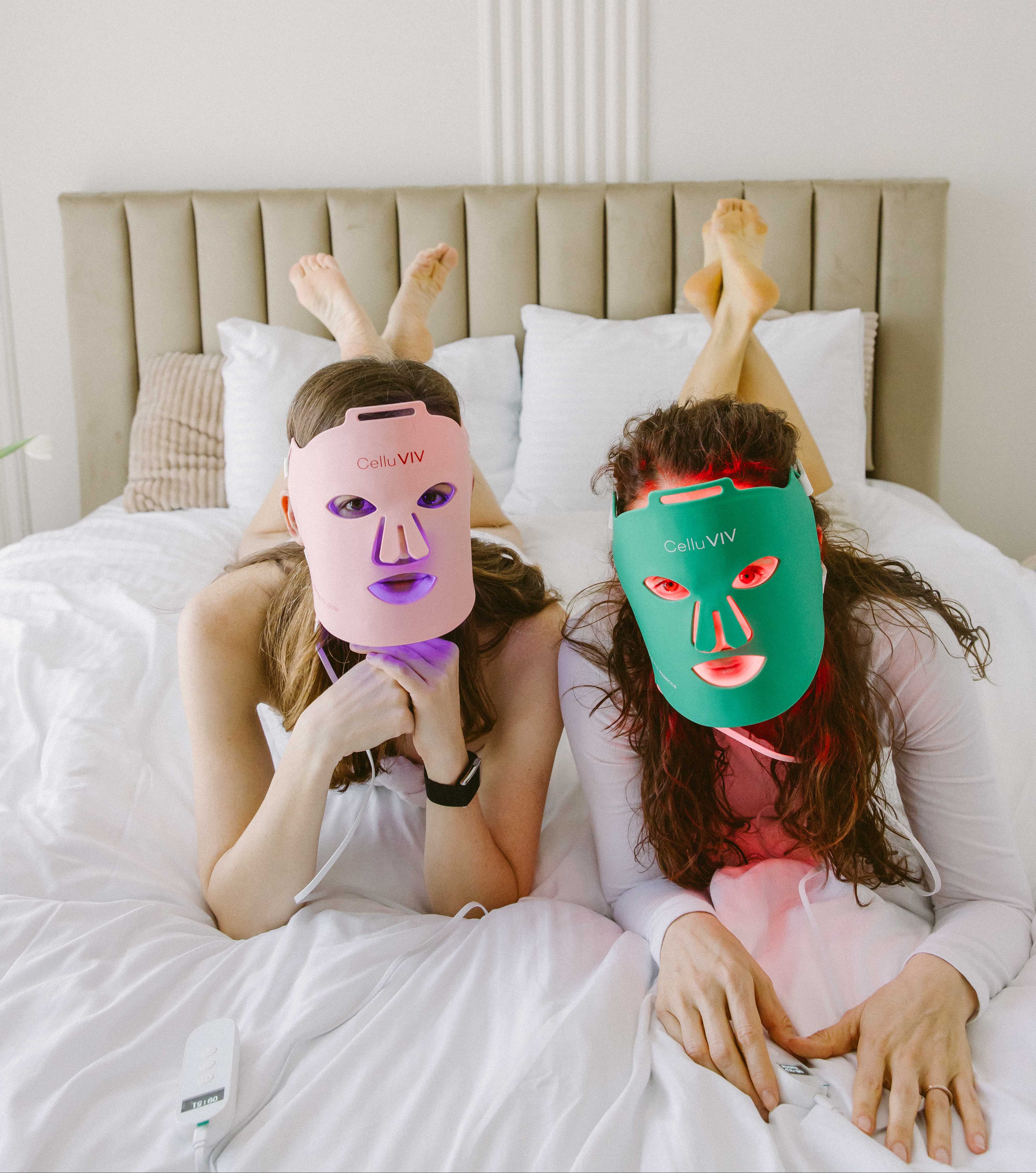 Two women lying on a bed wearing LED face masks in pink and emerald, doing LED light theraphy with blue light and red nd infrared light